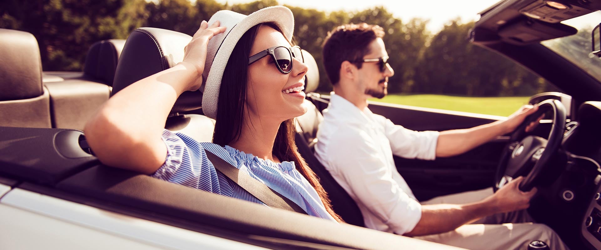 A woman and a man ride in a convertible car with the top down on a sunny day both are wearing sunglasses, and the woman is smiling.