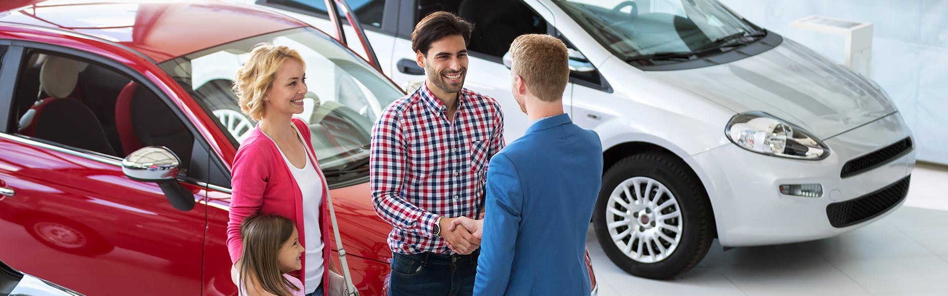 A smiling family shakes hands with a car salesman at a dealership, with red and silver cars in the background